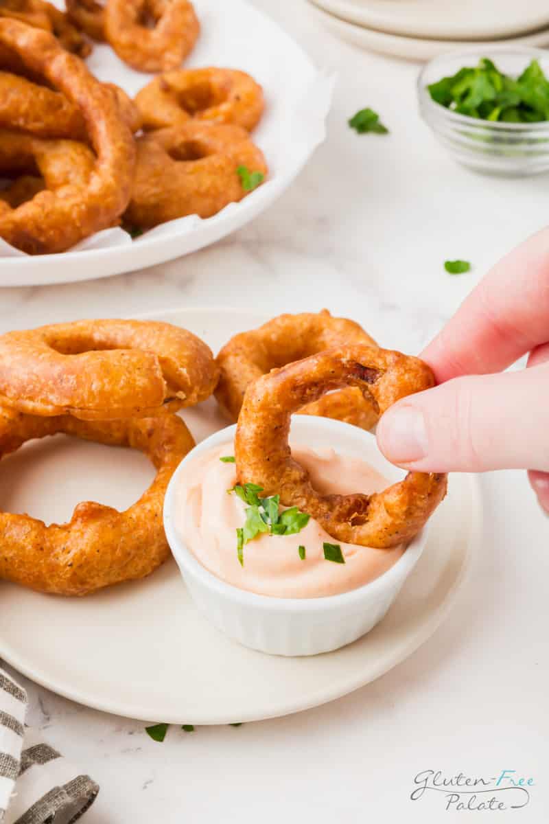 a small plate of onion rings and a cup of pinkish creamy dip. A hand is dipping one onion ring into the dip