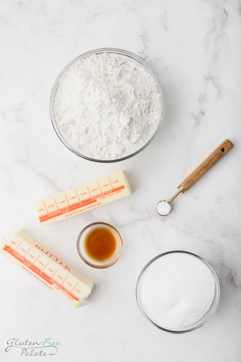 Ingredients for gluten-free shortbread cookies shown from the top down. Includes a bowl of flour, a bowl of sugar, a teaspoon of salt, a small bowl of vanilla, and a stick of butter