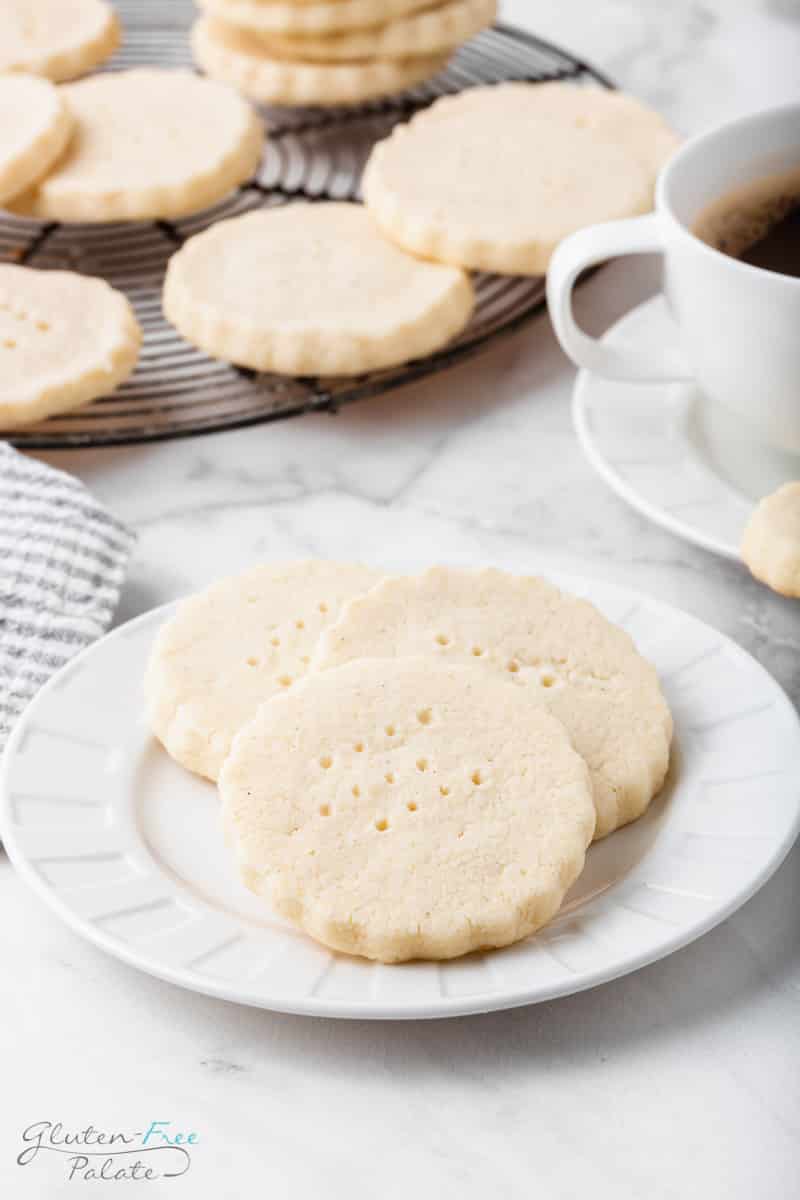 a white plate with three gluten-free shortbread cookies on it. The right is a teacup filled with coffee, and behind is a cooling rack filled with more cookies.