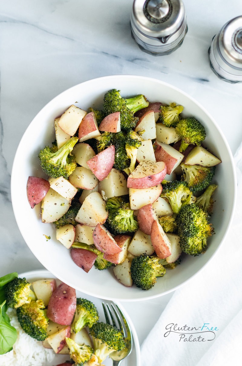 a large bowl of roasted potatoes and broccoli on a marble counter next to a plate with rice, potatoes, and broccoli and salt and pepper shakers