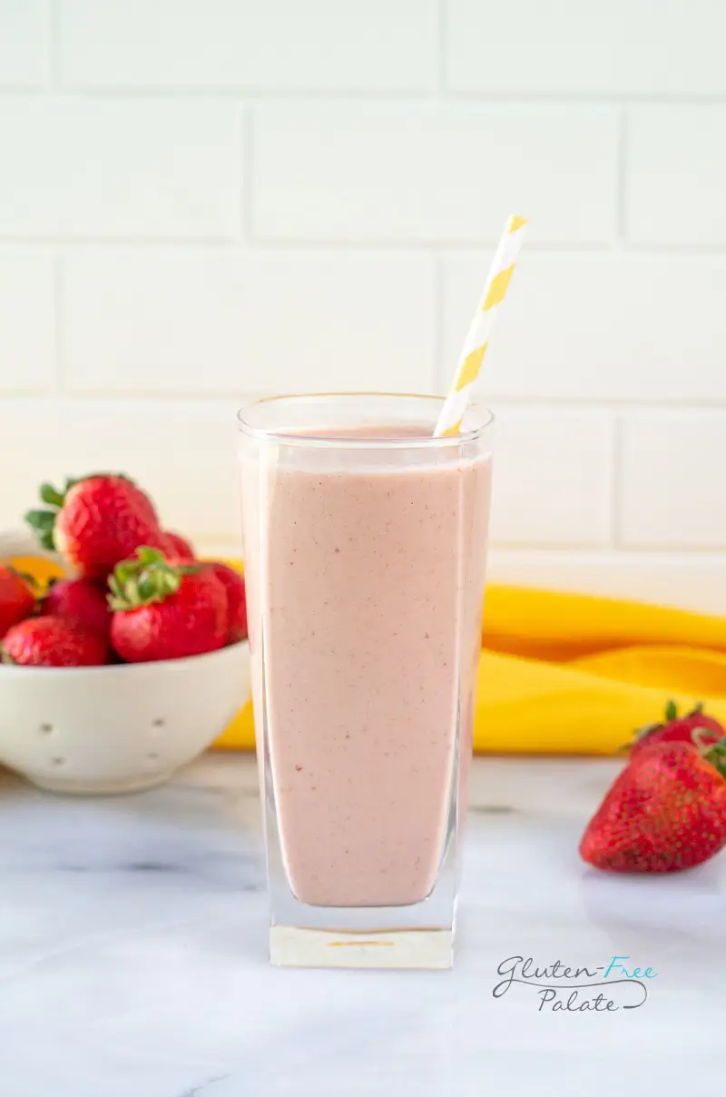 a tall glass filled with strawberry banana peanut butter smoothie, with a yellow and white striped straw. In the background is a bowl of strawberries and a yellow towel.