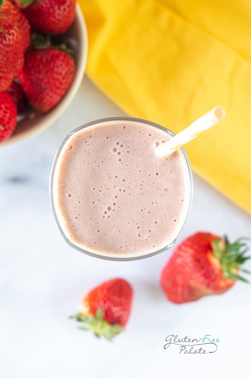 Top down view of a tall glass filled with strawberry banana peanut butter smoothie, with a yellow and white striped straw. In the background is a bowl of strawberries and a yellow towel.