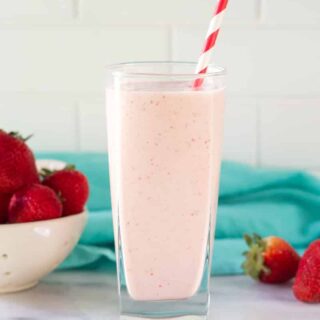 A tall glass filled with strawberry banana pineapple smoothie with a red and white striped straw. In the background is a bowl of strawberries and a teal kitchen towel.