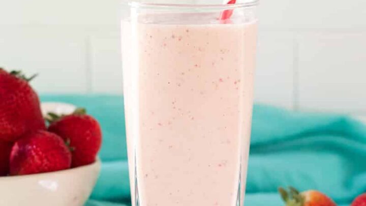 A tall glass filled with strawberry banana pineapple smoothie with a red and white striped straw. In the background is a bowl of strawberries and a teal kitchen towel.
