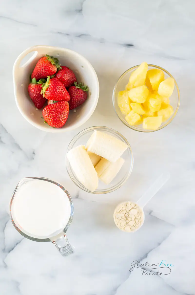 Ingredients for strawberry banana pineapple smoothie, all in separate bowls on a marble counter.