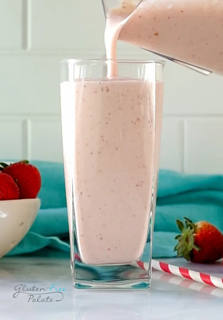 A tall glass being filled with strawberry banana pineapple smoothie from a pitcher. In the background is a bowl of strawberries and a teal kitchen towel.