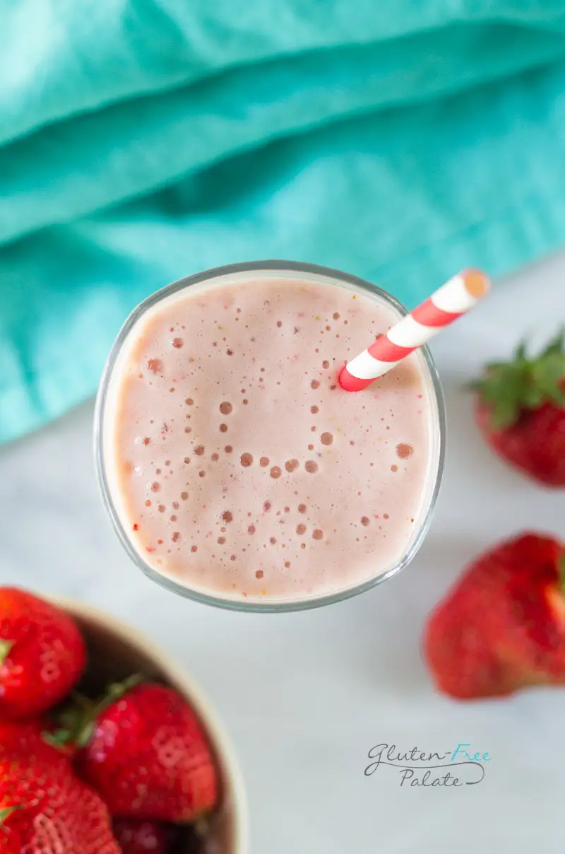 Top down view of a tall glass filled with strawberry banana pineapple smoothie with a red and white striped straw. In the background is a bowl of strawberries and a teal kitchen towel.