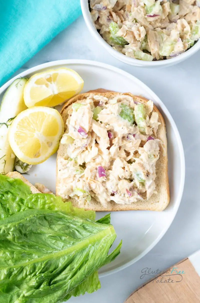 a white plate with a slice of white bread topped with tuna salad next to romaine lettuce leaves and quartered lemons.