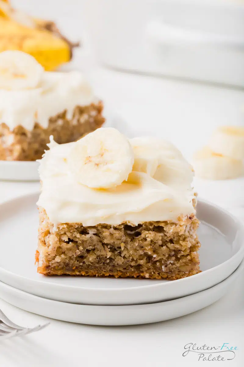 two white plates stacked, with a square piece of gluten-free banana cake with white frosting and a slice of banana on top.