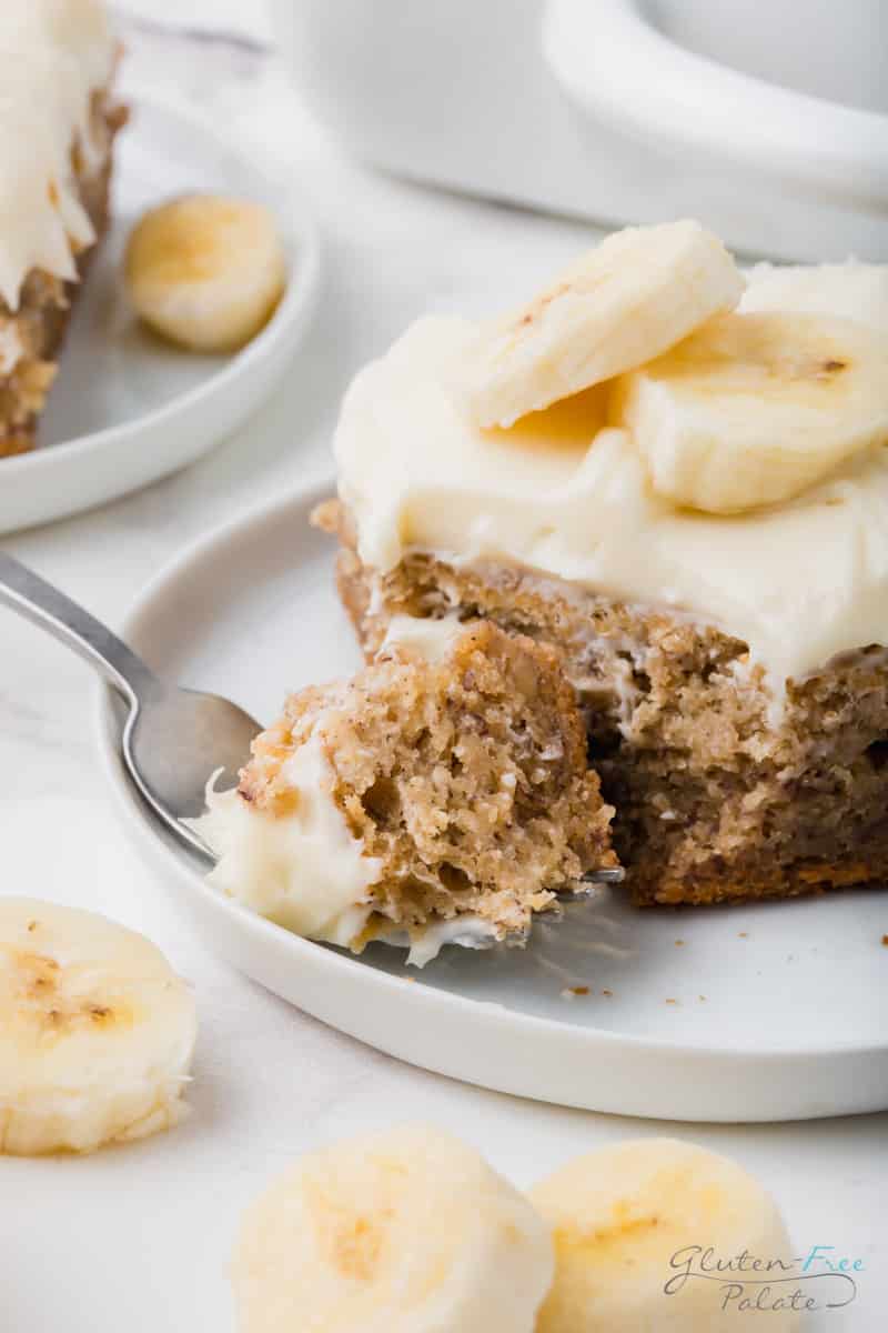 a plate with a slice of gluten-free banana cake being eaten with a fork. Cake and plate is garnished with sliced bananas.