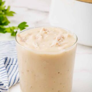 a clear, short glass filled with cream of chicken soup. A white pot , a bunch of parsley, and a blue striped kitchen towel are in the background.