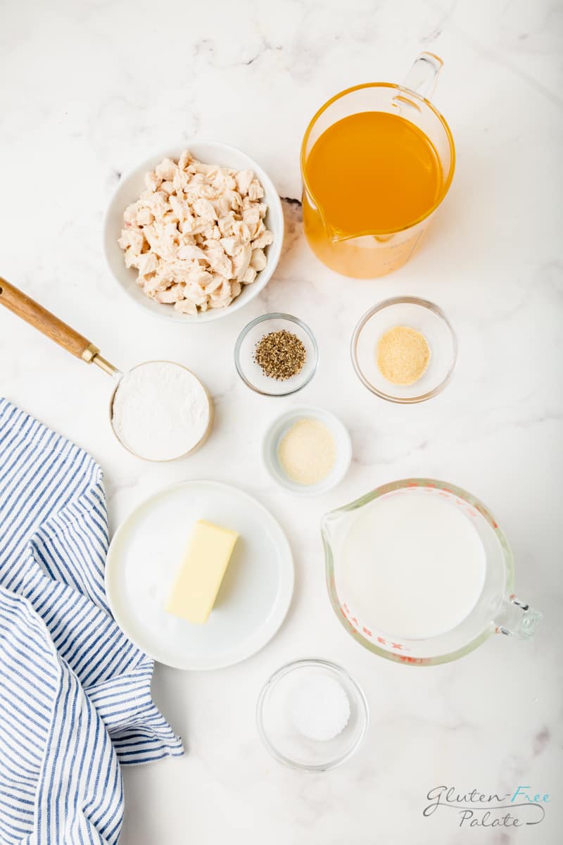 Top down view of the ingredients needed to make gluten free cream of chicken soup on a marble counter. Including a bowl of chicken, a pitcher of broth, a plate with butter, and small bowls of seasonings.