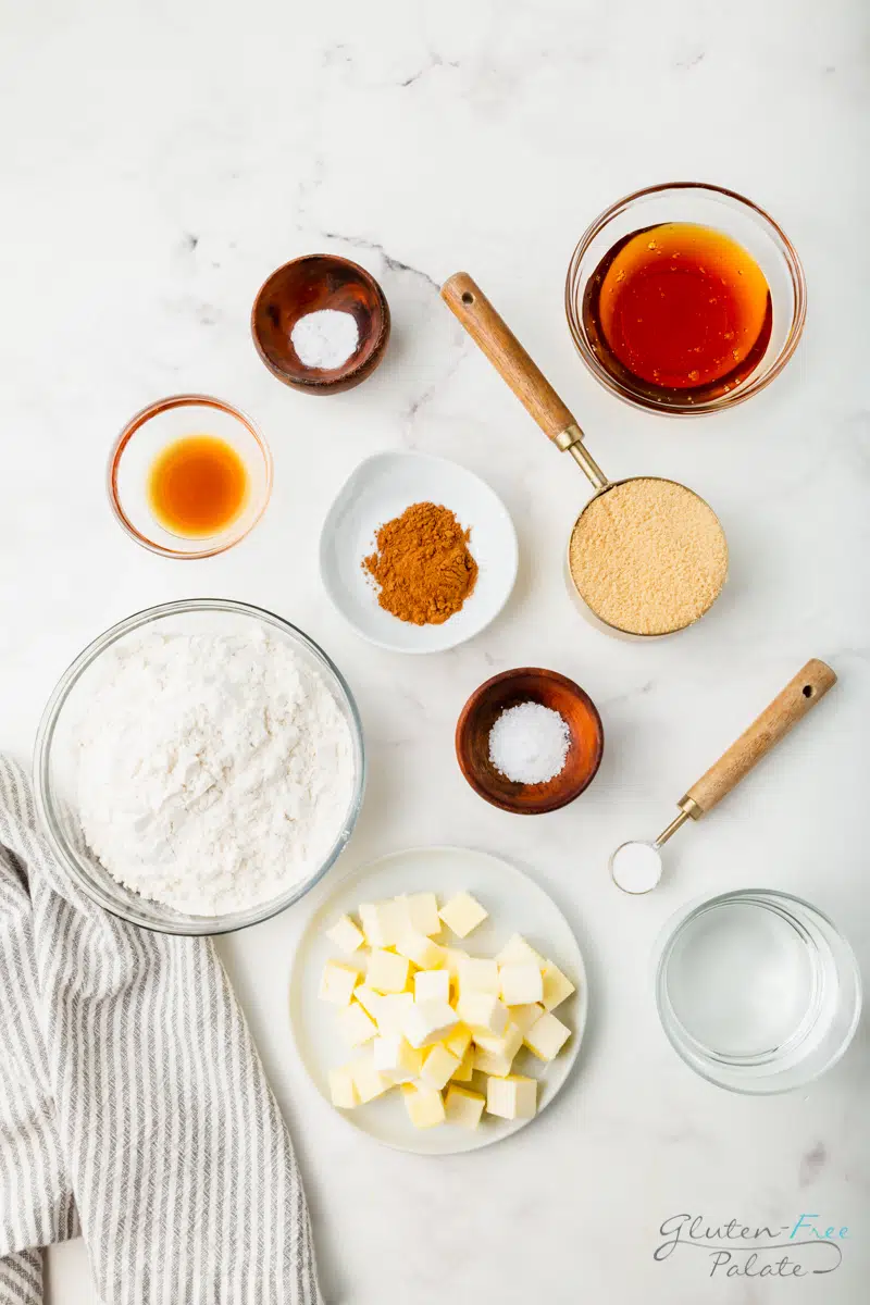 Top down view of the ingredients needed to make gluten-free graham crackers, all in separate bowls on a marble countertop. Includes a bowl of flour, diced butter on a plate, and other flavorings.