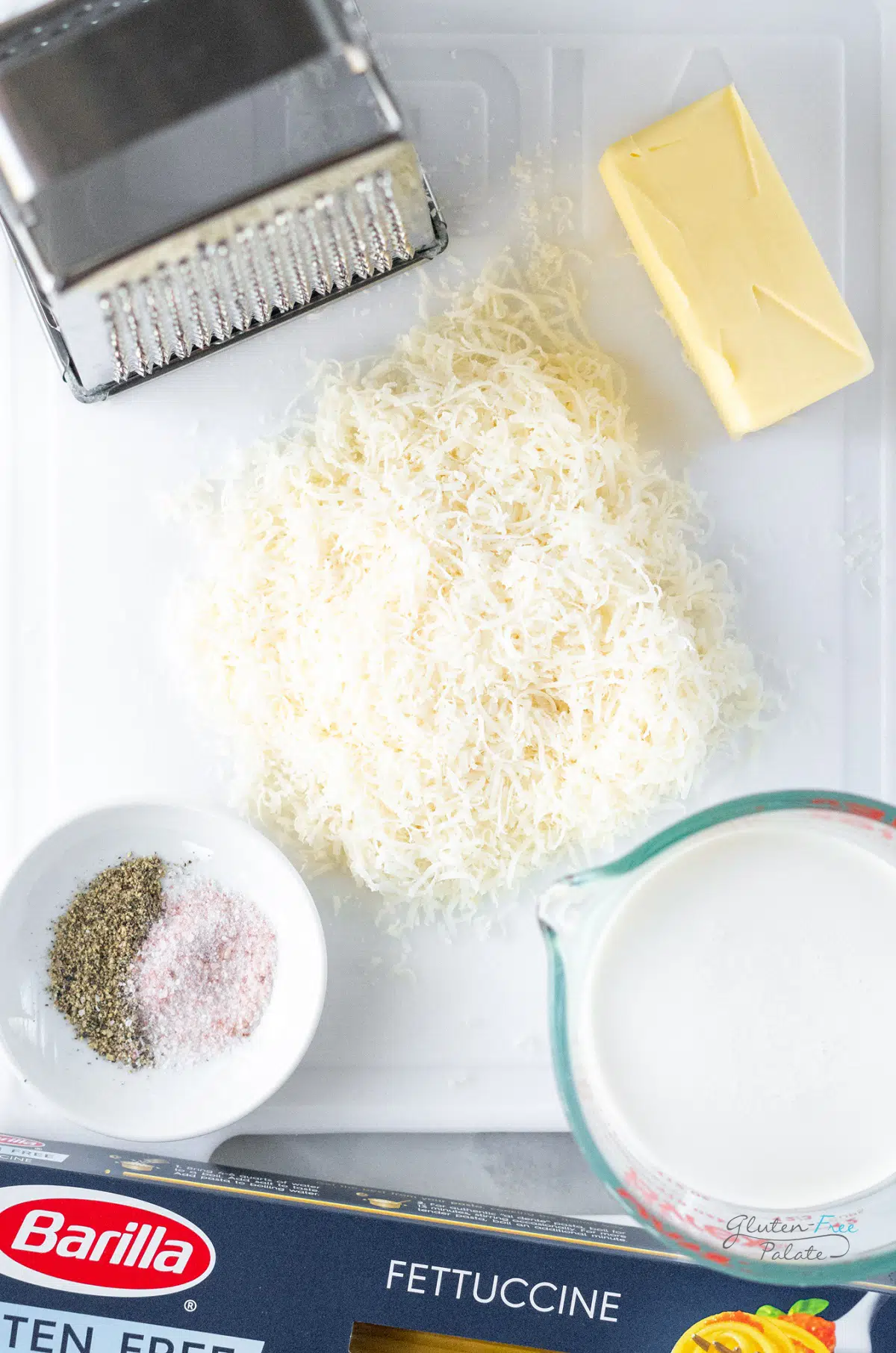 The ingredients for gluten free alfredo sauce on a white plastic cutting board with a box grater and a box of fettuccine