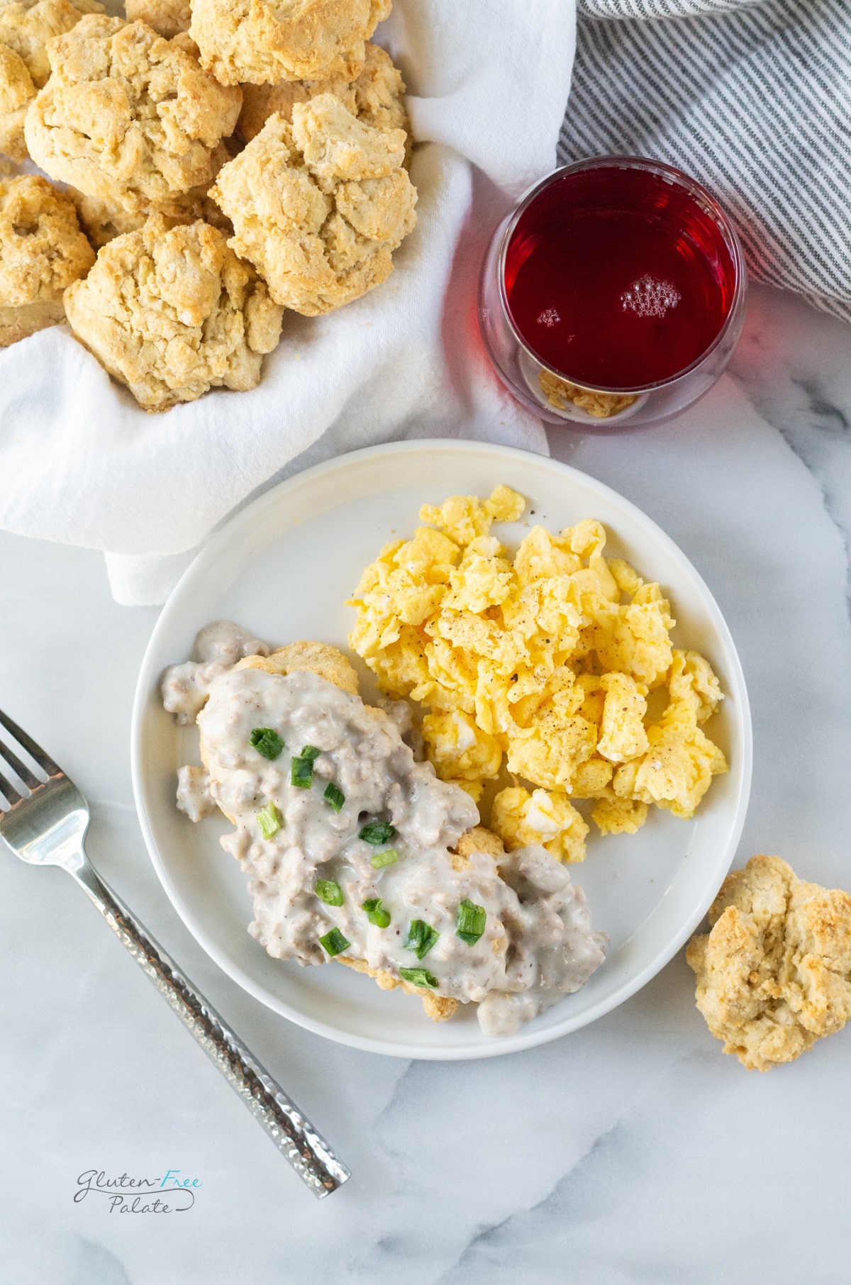 a breakfast plate of scrambled eggs, biscuits topped with gravy. A silver fork is on the left, a basket of fresh gluten free biscuits and a glass of cranberry juice is above the plate.