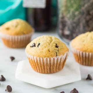 a perfectly domed chocolate chip muffin on a white hexagon pedastal. In the background are two more muffins. Chocolate chips are scattered on the counter