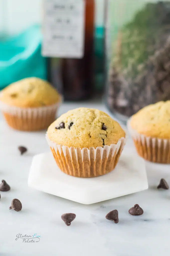 a perfectly domed chocolate chip muffin on a white hexagon pedastal. In the background are two more muffins. Chocolate chips are scattered on the counter