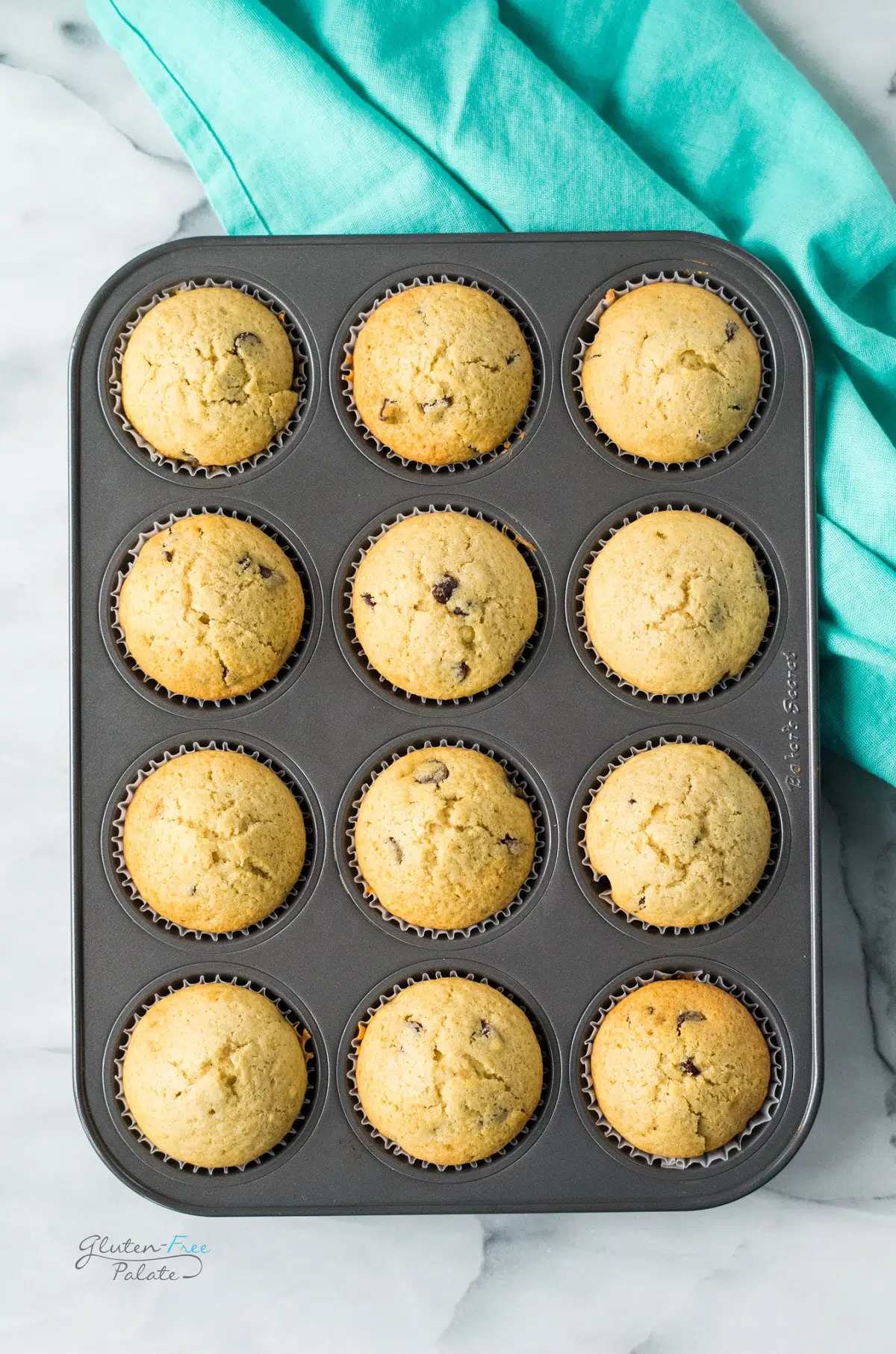 View from above of 12 gluten-free chocolate chip muffins in a muffin pan. a teal towel is underneath the pan.