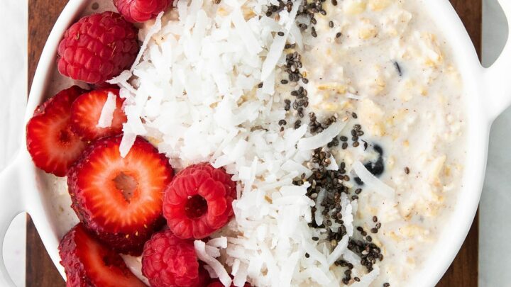 a white bowl with handles, filled with creamy overnight oats topped with seeds, coconut, and fresh berries.