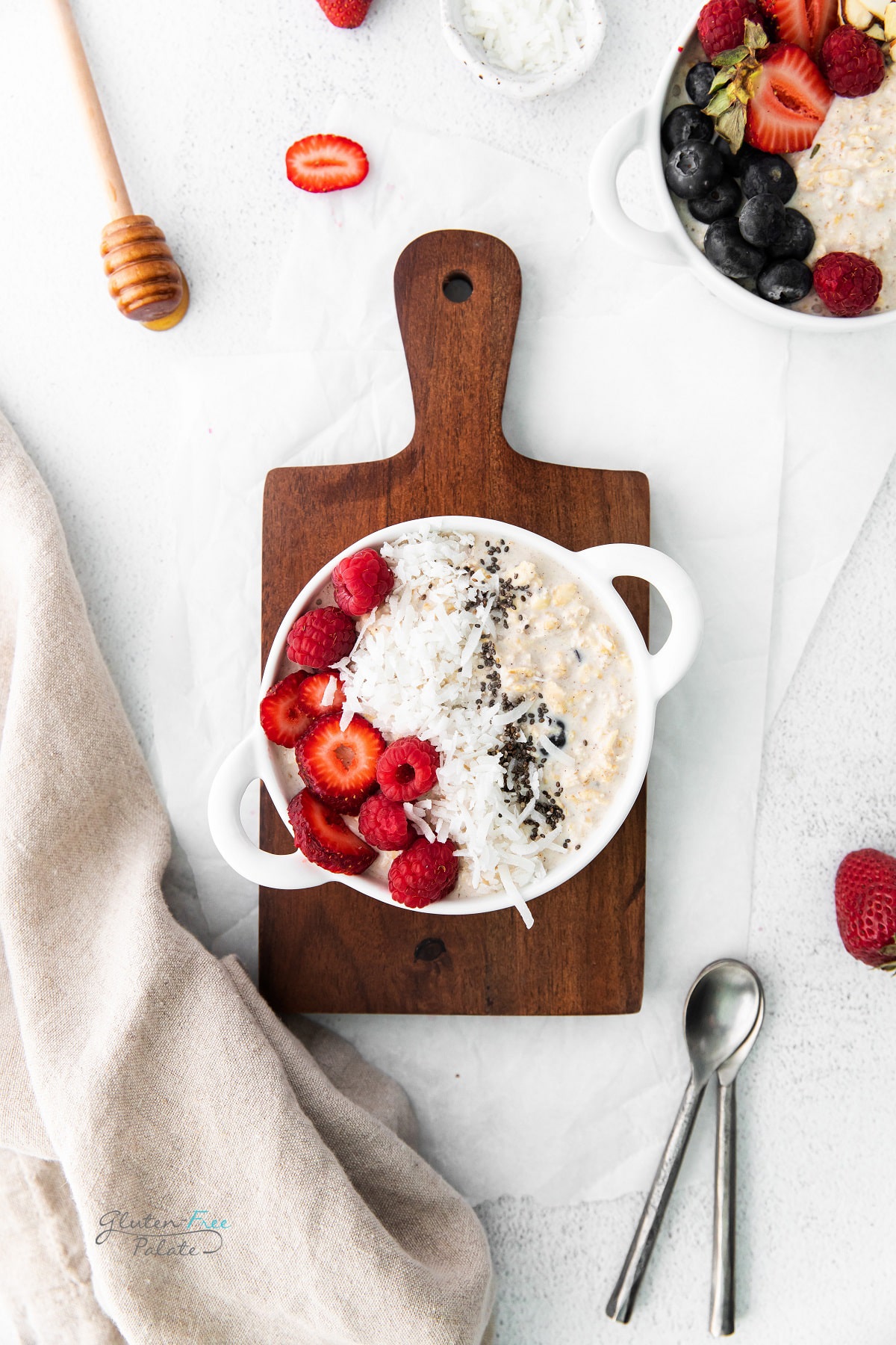a wooden cutting board on a table, with a bowl of overnight oats with berries, coconut toppings. another bowl is nearby with blueberries and strawberries in it. A honey dipper is on the table as well.