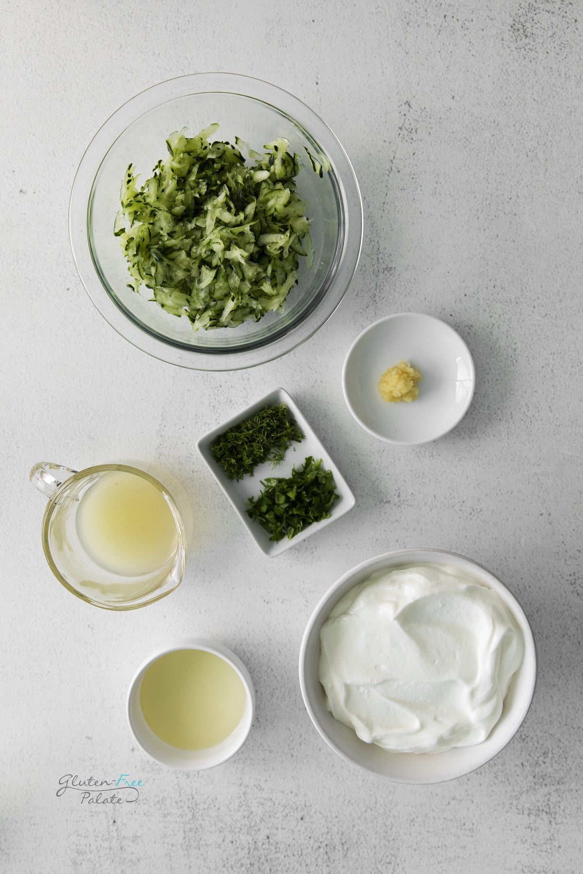 The ingredients for homemade Tzatziki dip, measured into separate small bowls on a concrete counter, viewed from above