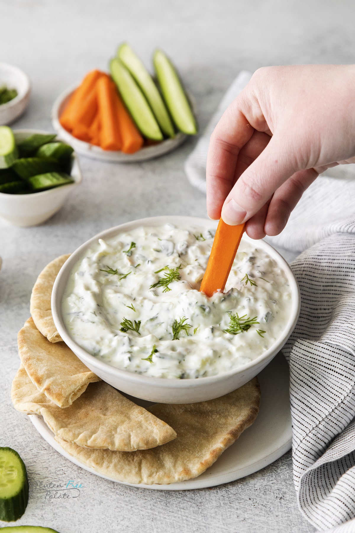 a white bowl of homemade tzatziki on a plate of pita bread. A hand is dipping a carrot stick into the dip