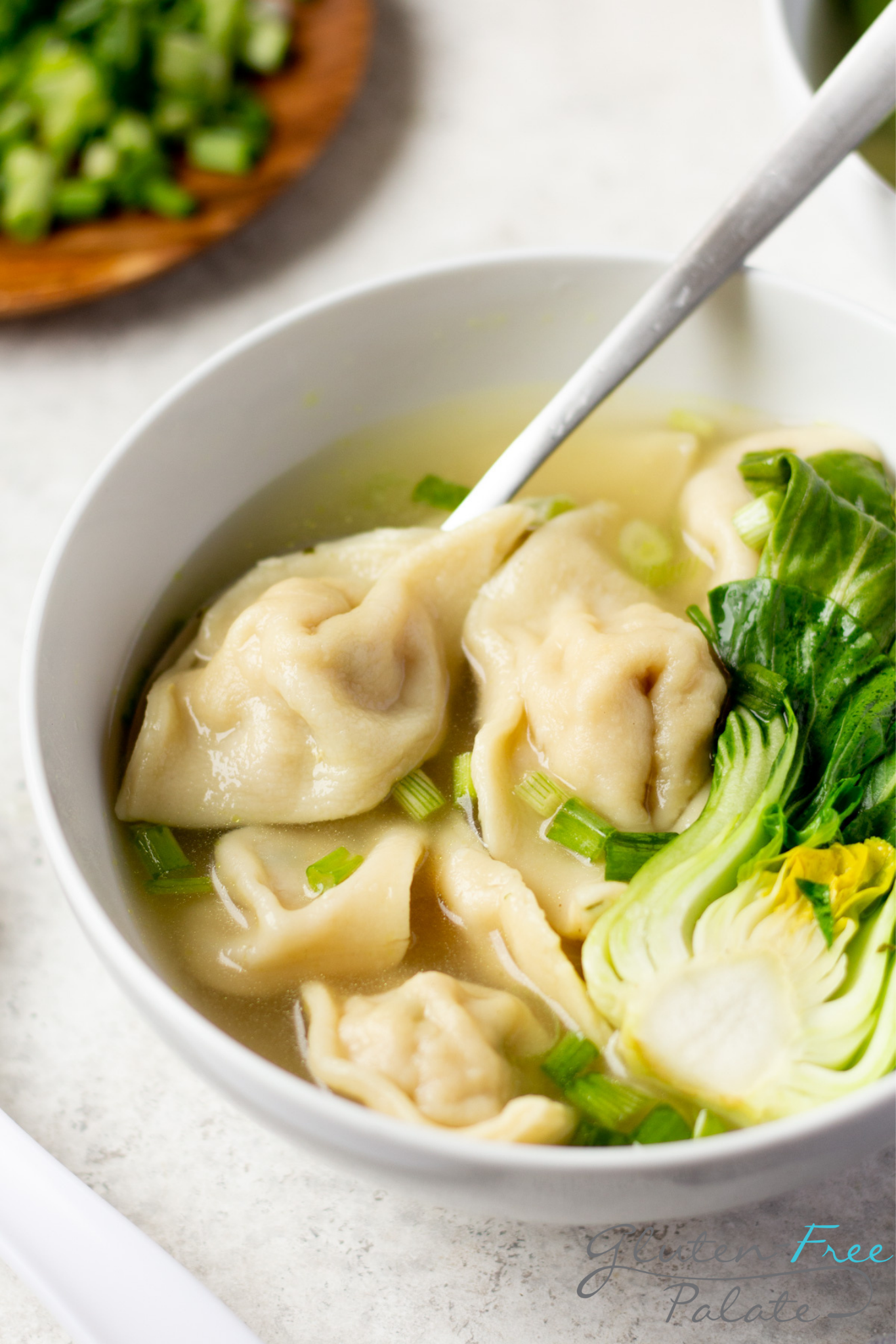 a bowl of wonton soup with baby Bok choi. A spoon is lifting a gluten-free wonton wrapper.