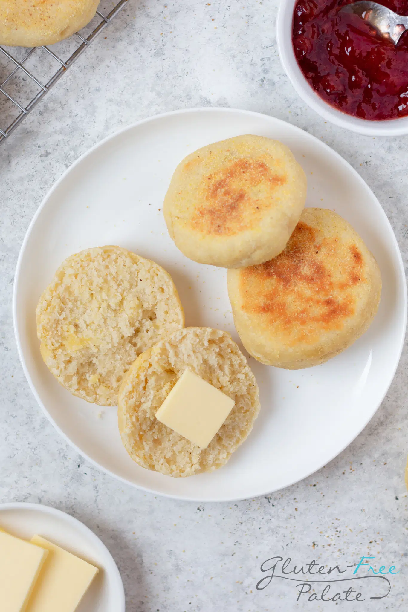 a round white plate with three gluten-free english muffins on it. One has been split in half and a pat of butter is on one side.