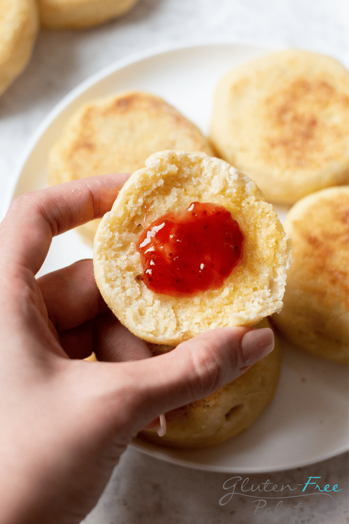 a feminine hand holding a gluten free english muffin half topped with butter and strawberry jam.