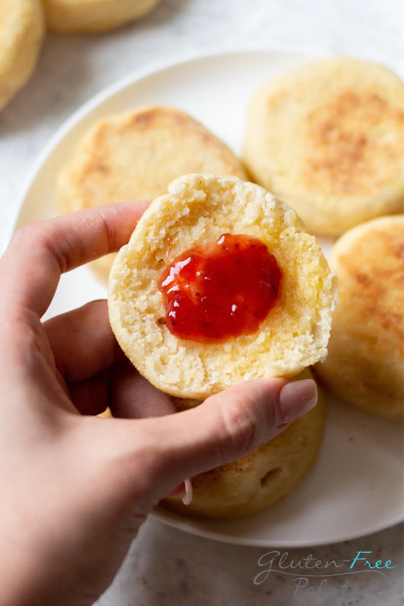 a feminine hand holding a gluten-free english muffin half topped with butter and strawberry jam.