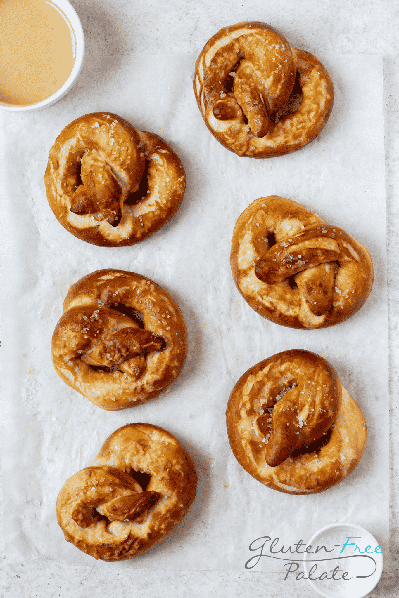 a parchment lined pan with six large homemade gluten-free pretzels on it. A cup of honey mustard is on the corner of the pan. 
