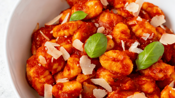 a bowl of gluten free gnocchi in tomato sauce garnished with parmesan cheese and fresh basil leaves