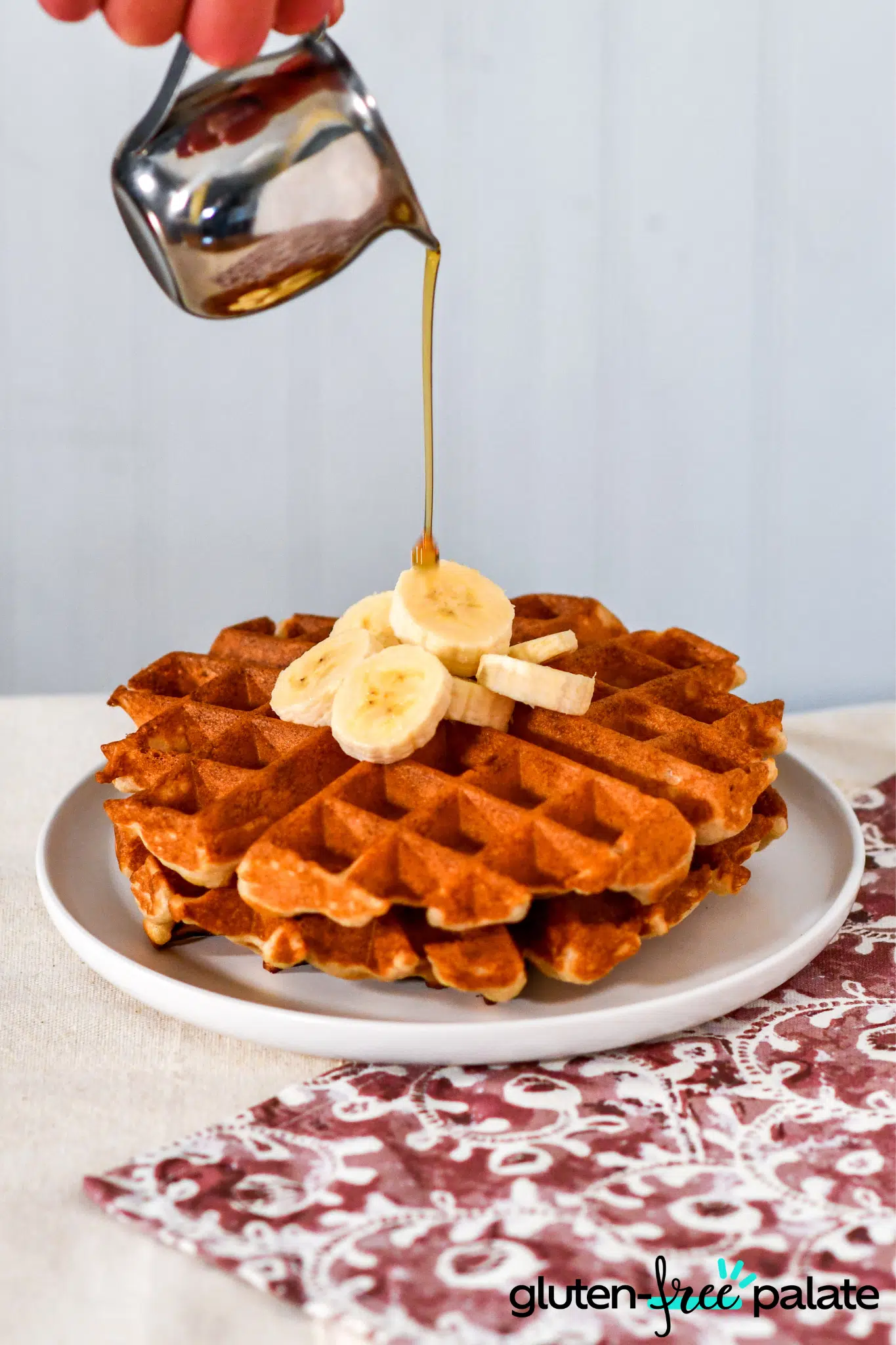 Gluten-free Maple Waffles with banana on a white plate with syrup being poured on it.