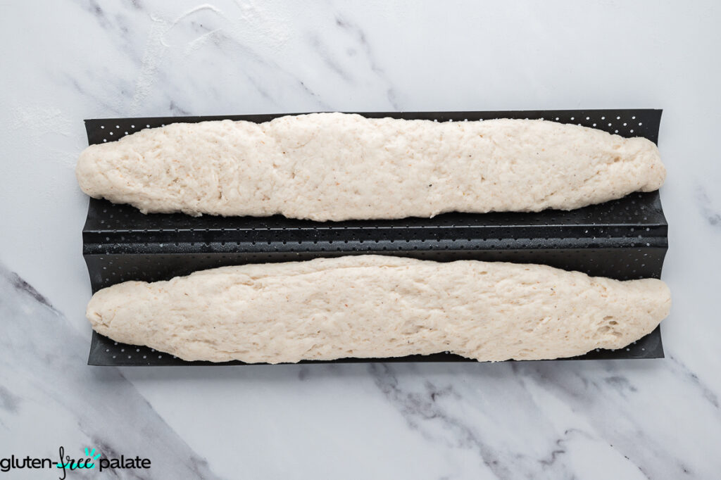 Two shaped gluten-free baguette loaves placed in a perforated baguette pan, ready to proof before baking.