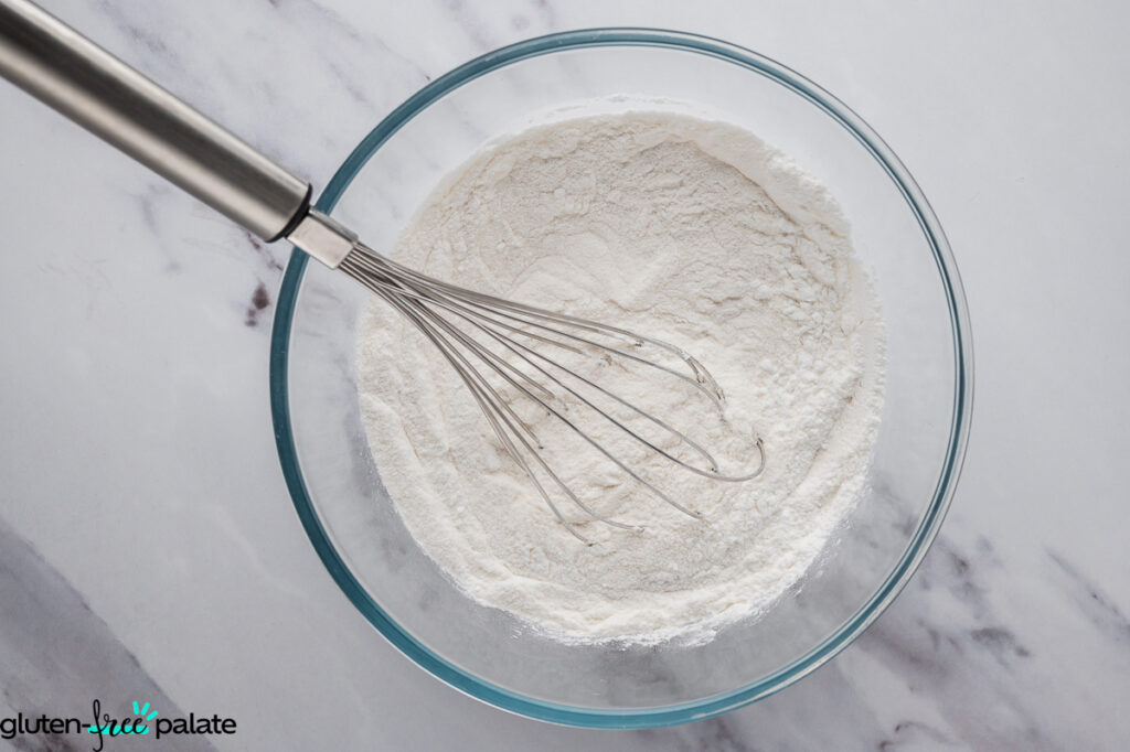 clear glass bowl filled with gluten-free flour blend, with a stainless steel whisk resting inside.