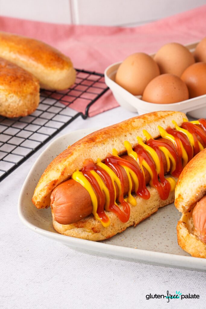 Gluten-free hot dog buns on a plate. Topped with ketchup and mustard. Photo by Gluten-Free Palate.