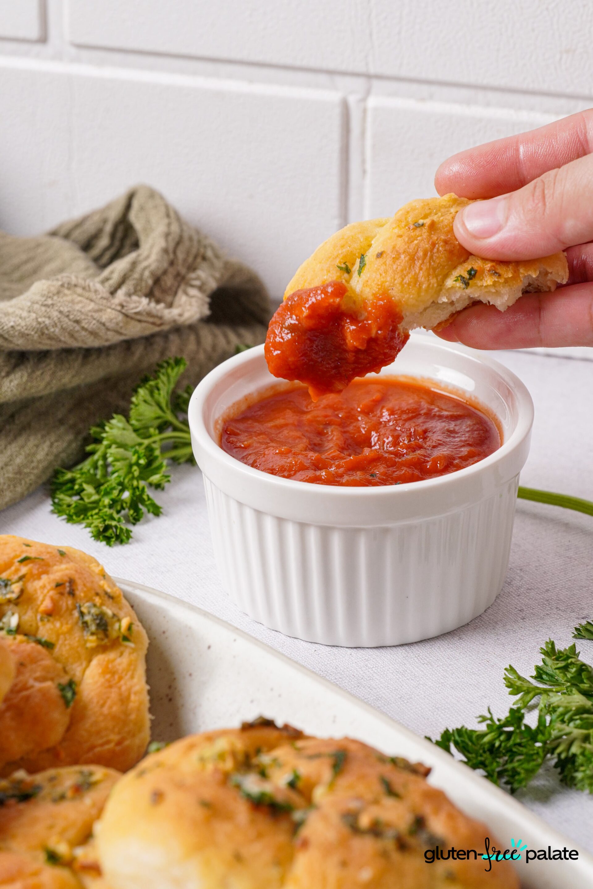 Gluten-free garlic knots on a white plate with toppings; one is dipped into a sauce.