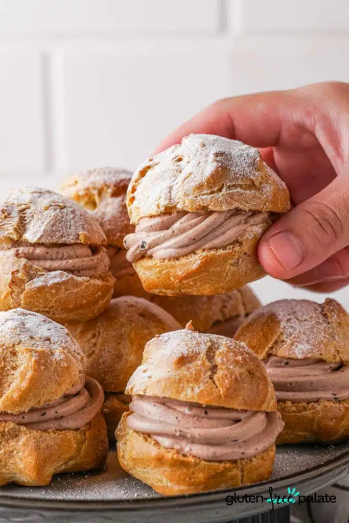 Gluten-Free Choux Pastry made into cream puffs on a cake stand with icing sugar on top and a hand holding one.
