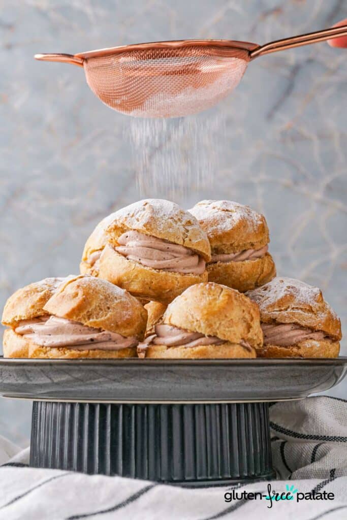 Gluten-Free Choux Pastry made into cream puffs on a cake stand with icing sugar being sprinkled on top.