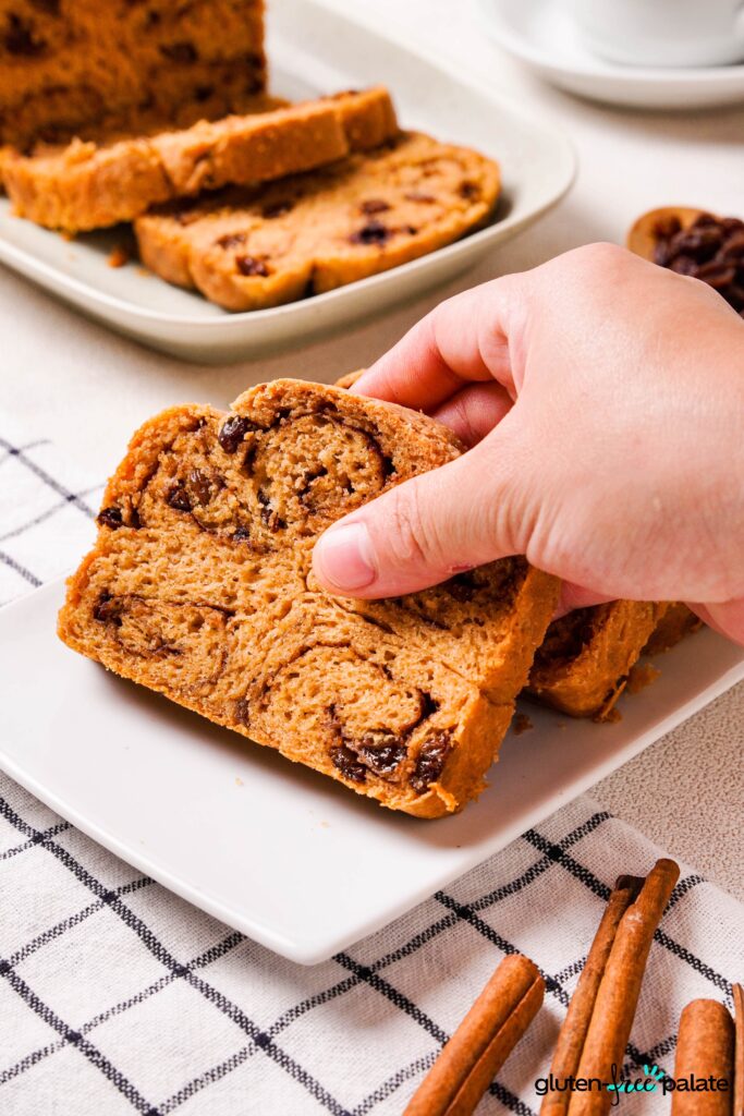 Gluten-Free Cinnamon Raisin Bread on a white plate sliced being picked up by a hand.