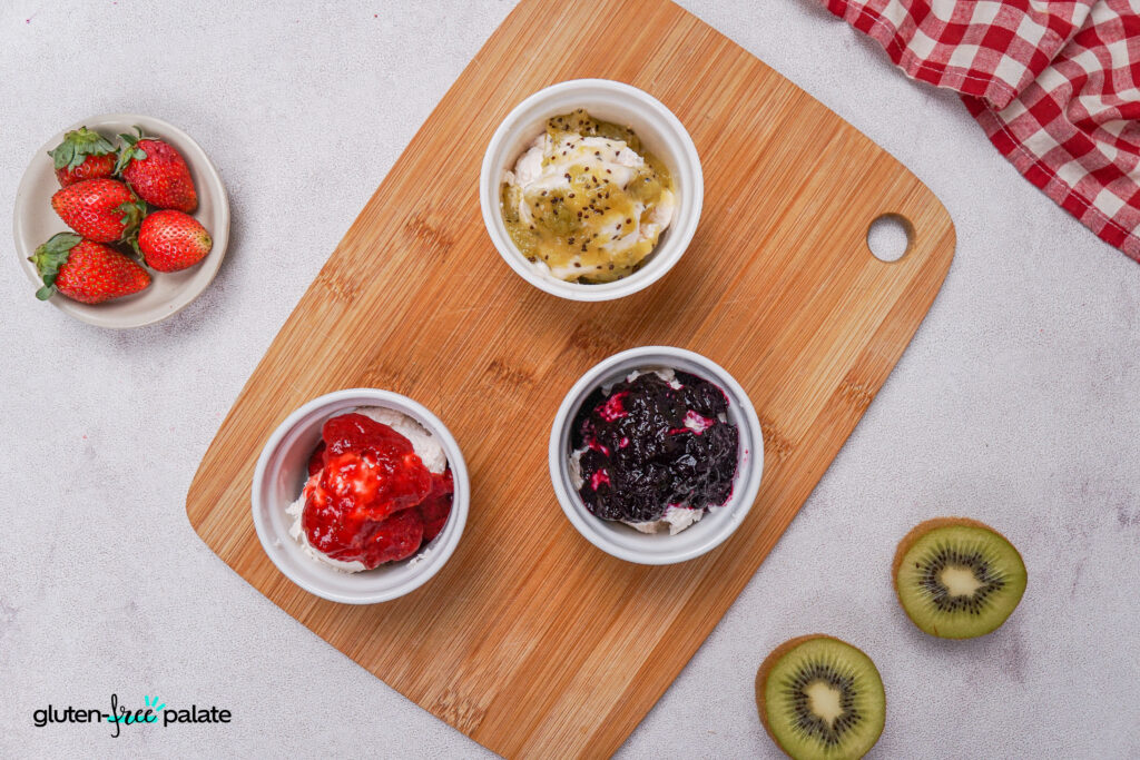 Three small ramekins of vanilla ice cream topped with different fruit compotes—strawberry, kiwi, and berry—arranged on a wooden board with a bowl of fresh strawberries and sliced kiwi nearby.