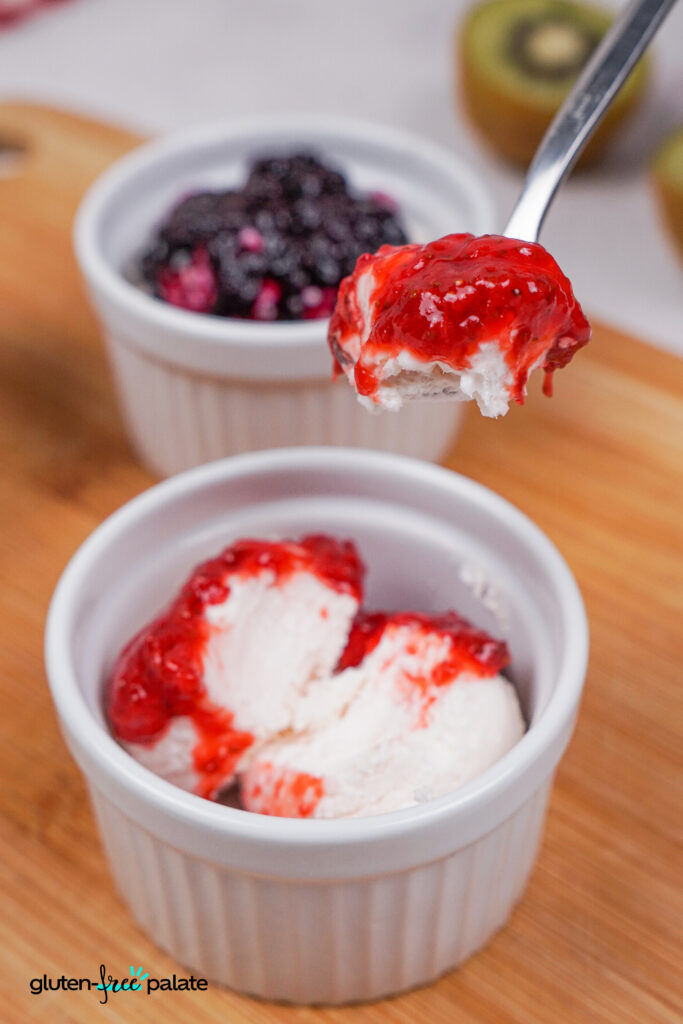 A spoonful of vanilla ice cream topped with bright red strawberry compote, lifted above a small white ramekin of the same dessert. In the background, a second ramekin with dark berry compote is slightly blurred.
