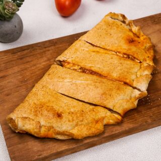 A gluten-free calzone sliced into portions, showing its golden crust, placed on a wooden cutting board with fresh tomatoes nearby. Photo by Gluten-Free Palate.