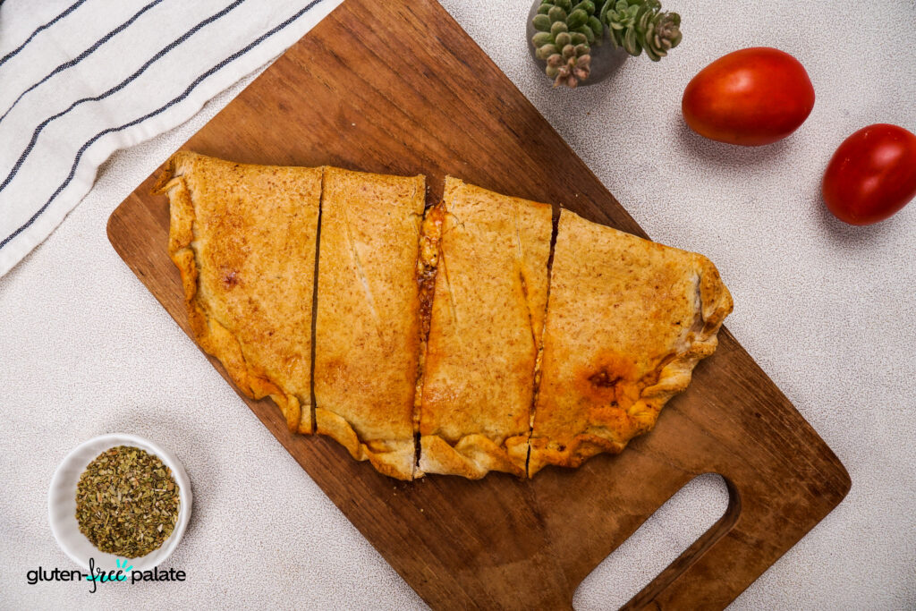 A top-down view of a gluten-free calzone on a cutting board, sliced into portions, surrounded by fresh tomatoes and a bowl of Italian seasoning. Photo by Gluten-Free Palate.