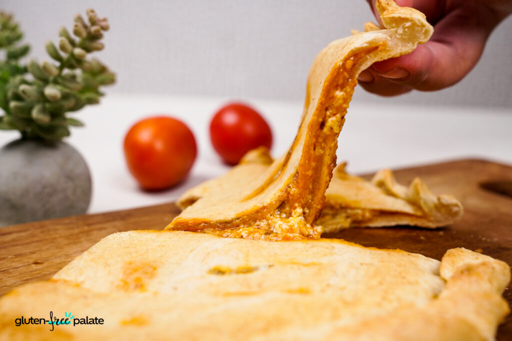 A hand lifting a slice of gluten-free calzone, revealing a gooey, cheesy interior with marinara sauce. Photo by Gluten-Free Palate.