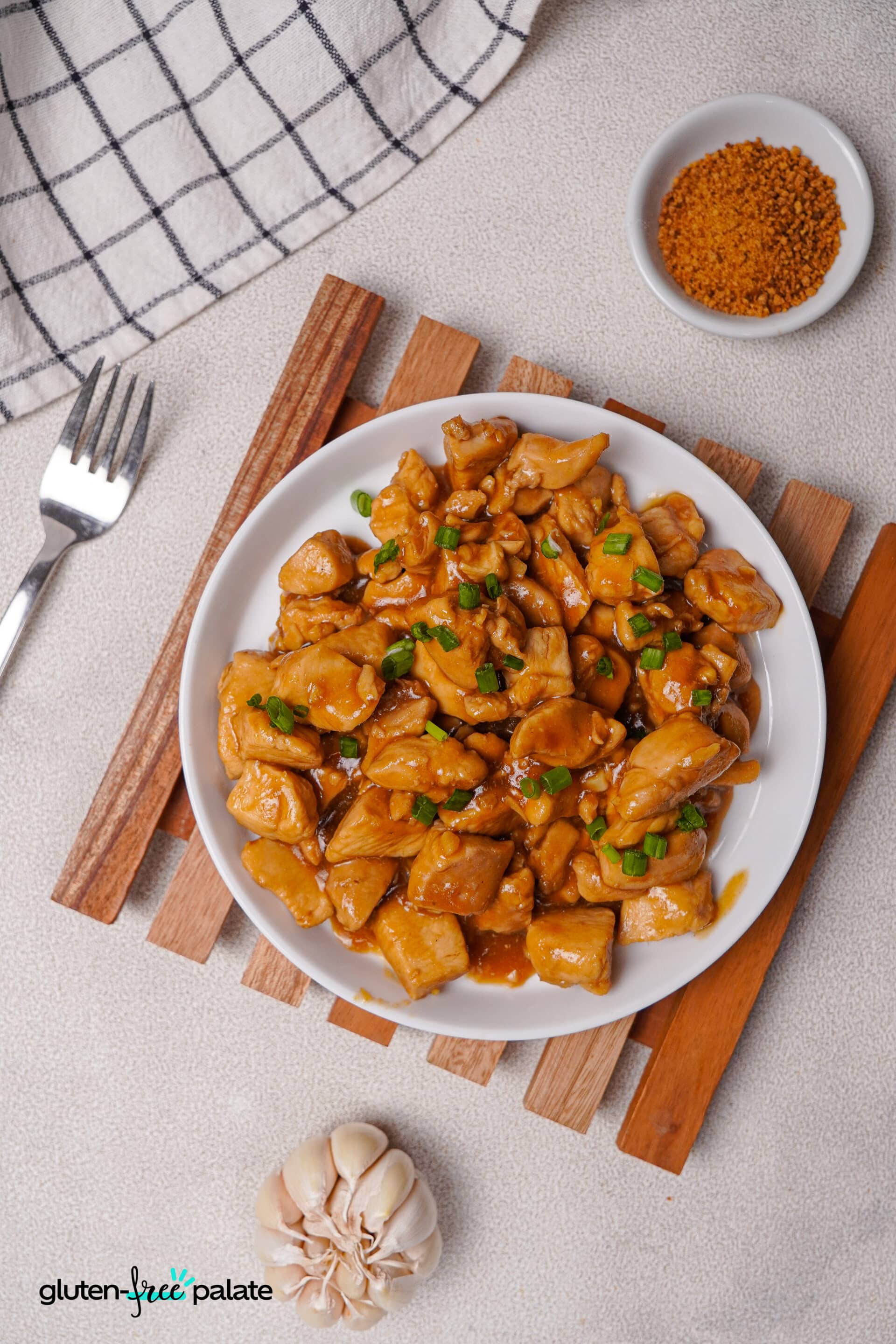 A table setup featuring a plate of teriyaki chicken with a fork, a garlic bulb, and a small bowl of brown sugar, all placed on a textured countertop. Photo by Gluten-Free Palate.