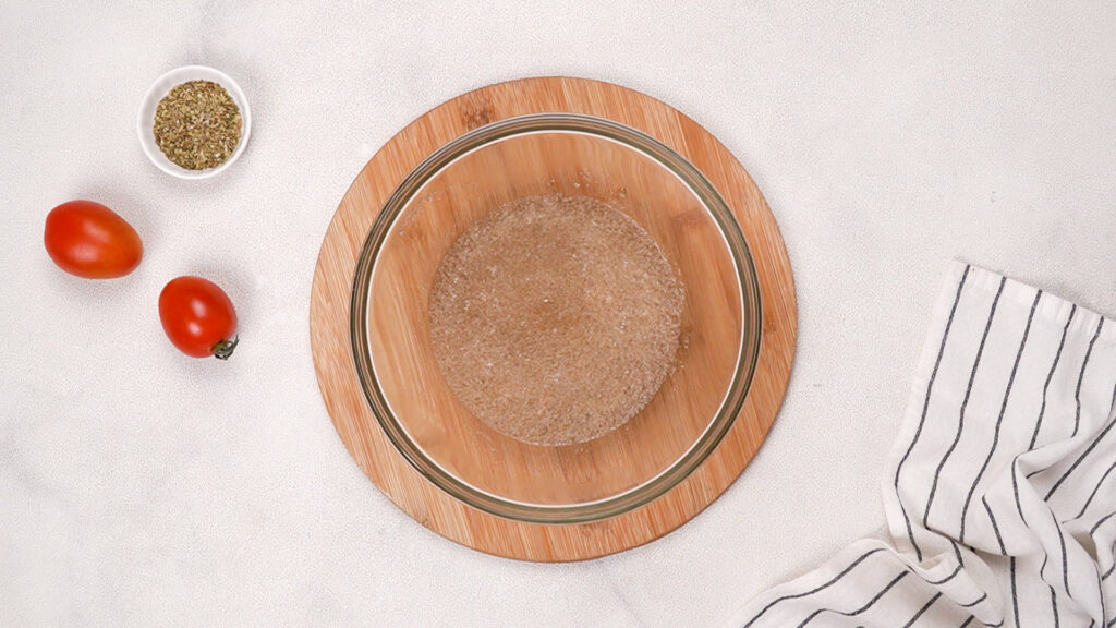 A glass bowl with psyllium husk and water forming a gel, placed on a wooden board, with fresh tomatoes, a bowl of Italian seasoning, and a striped towel. Photo by Gluten-Free Palate.