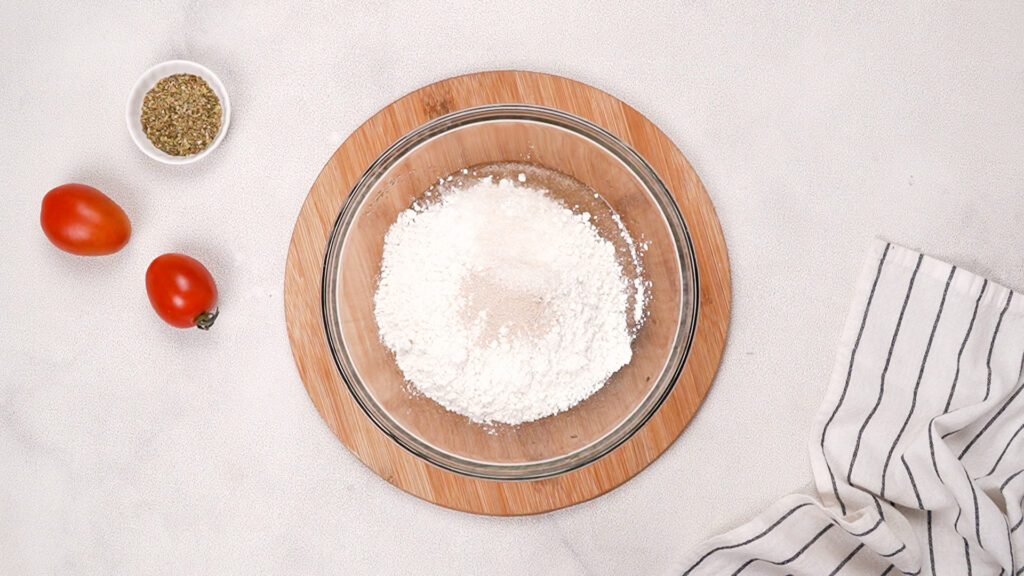 A glass bowl filled with gluten-free flour, yeast, and other dry ingredients on a wooden board, surrounded by fresh tomatoes, seasoning, and a striped towel. Photo by Gluten-Free Palate.