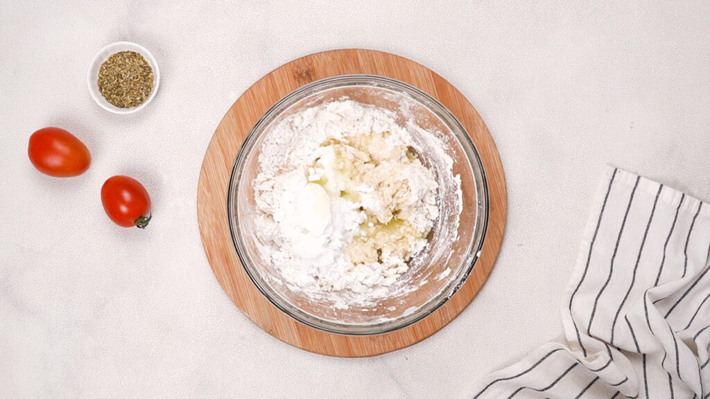 A glass bowl showing gluten-free flour mixture combined with olive oil and psyllium gel, ready to be mixed into a dough, with tomatoes and seasoning nearby. Photo by Gluten-Free Palate.