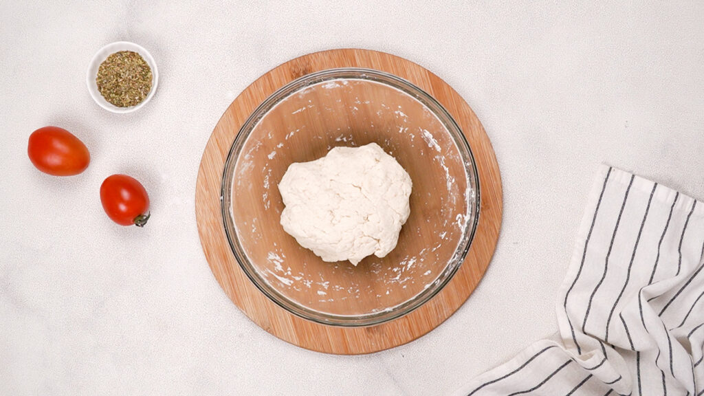 A glass bowl containing gluten-free dough on a wooden board, with fresh tomatoes and a bowl of seasoning in the background. Photo by Gluten-Free Palate.